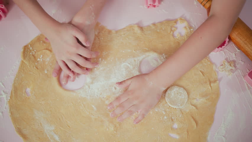 Overhead view of kids pressing pink shape cutters into dough while one rubs flour across surface and quickly switches to cutting another shape, capturing playful baking moment on pink floured table