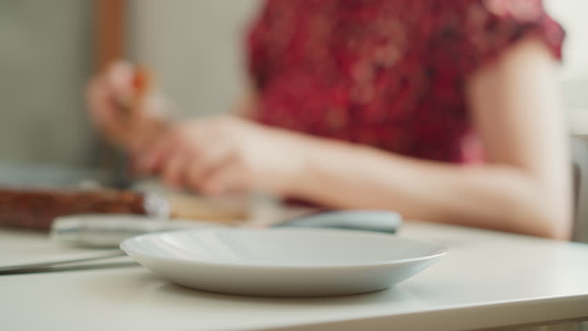 Young woman gently drops peeled sausage casing into white bowl on bright kitchen table, motion blur capturing fluid movement and casual meal prep under soft window light