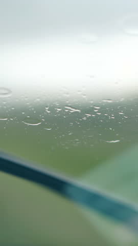 close view black metal wiper cleans raindrops from car windshield against blurred meadow and sky