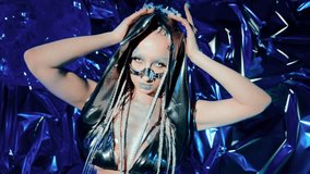 A woman in a chrome hood with an icy gaze and a facial accessory near her nose poses against a blue foil background. Her glitter makeup and braids highlight a futuristic fantasy sci-fi portrait. - Powered by Shutterstock - Get 15% off with code: PIKWIZARD15
