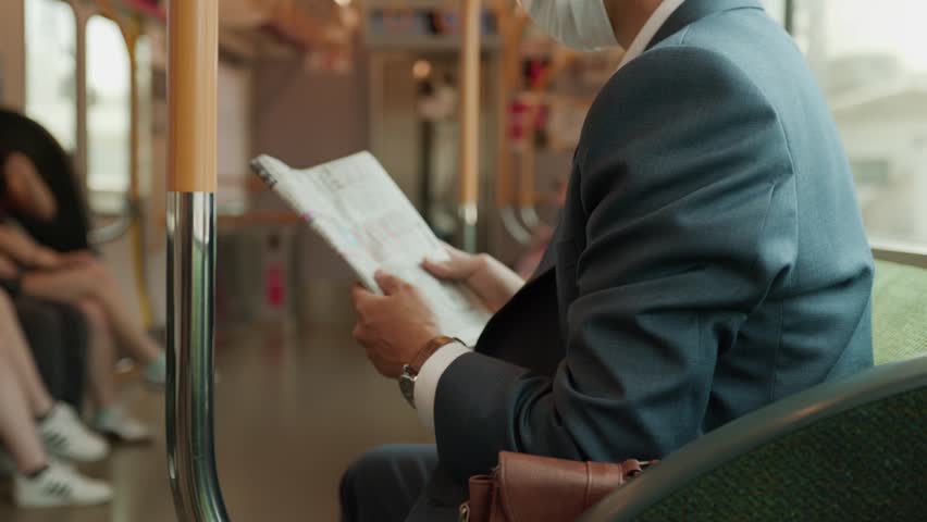 Young businessman in suit and mask reads a newspaper while sitting on a subway train, symbolizing urban commuting, information update, and health safety.