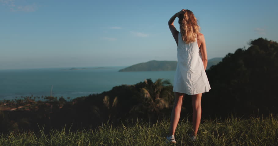 Woman in white dress stands on hill ocean and a small island in the distance. Girl relax tropical island , enjoy wild nature landscape. Outdoor lifestyle travel, summer holiday vacation, slow motion