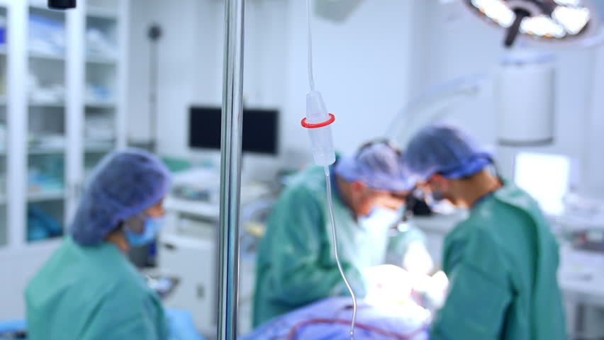 Drop counter on the stand in the surgery room. Medical team operating a patient at blurred backdrop.