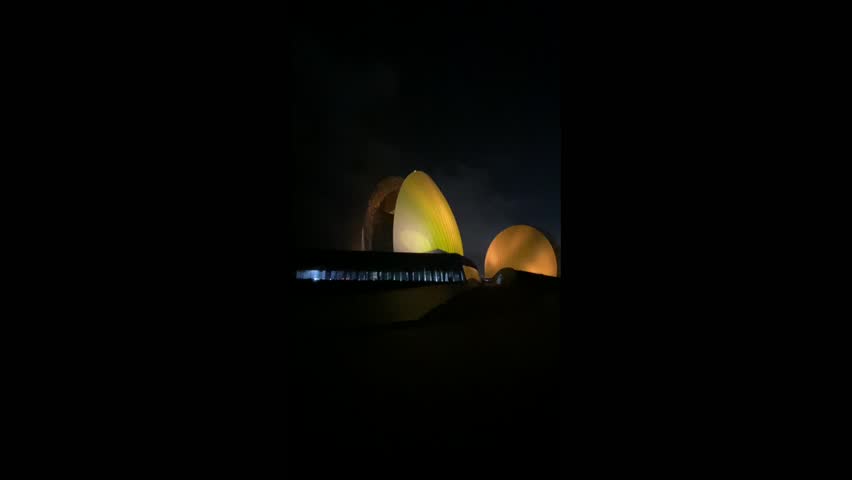 The grand Zhuhai Opera House is illuminated with golden yellow light at night. Its unique seashell-like forms gleam at the edge of the dark water. The night sky is partly cloudy with faint city lights