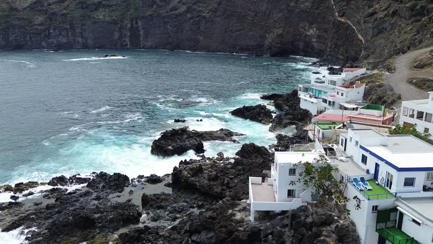 Aerial footage of a small coastal village with white houses built along the rocky shoreline of Tenerife island, Canary Islands, Spain. Blue ocean waves crashing against the volcanic rocks.