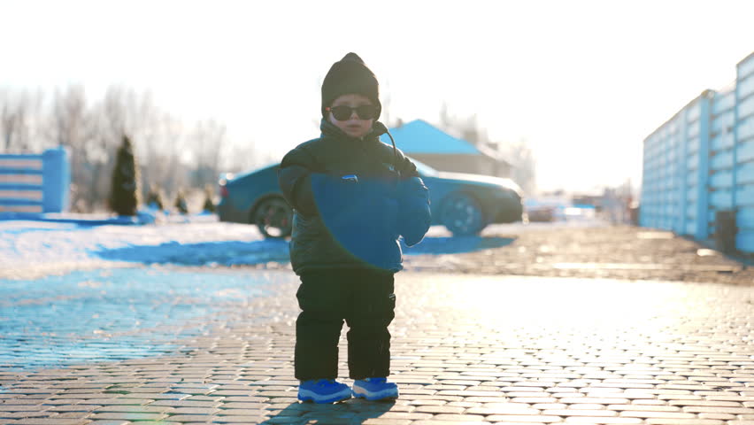 Cute fashionable baby boy wearing black cap, blazer, pants and blue shoes. Kid stands outdoors holding key from car at backdrop.