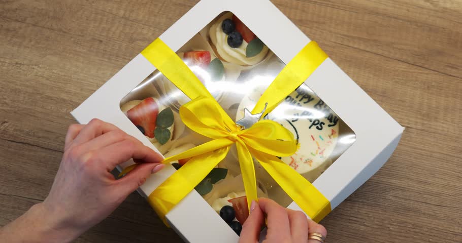 Birthday girl unties bright ribbon to reveal square box containing cakes. Novel sweet treat in special packaging lifts mood and helps to satisfy hunger