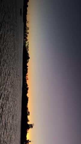 City skyline at sunset with glowing orange horizon and calm water in the foreground. Silhouetted buildings and peaceful ripples create a tranquil, cinematic urban landscape ideal for travel