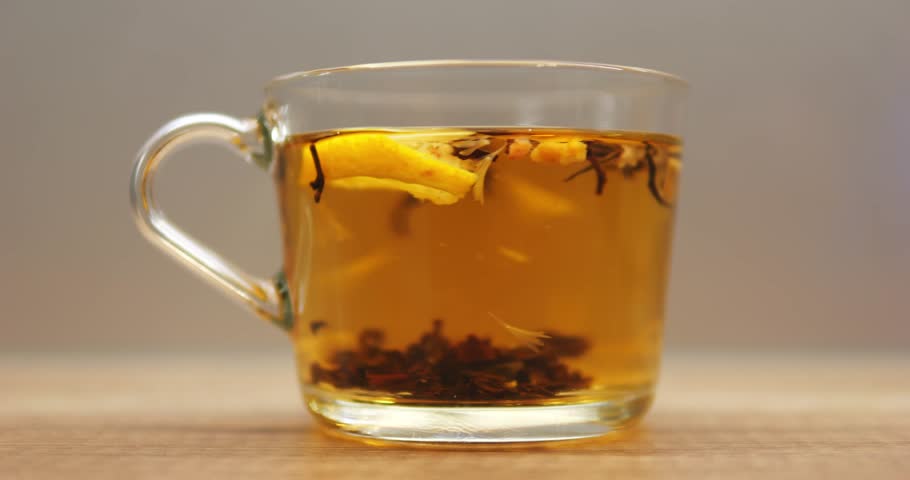 Tea leaves and fresh lemon slice rotate in transparent mug with hot water. Process of brewing herbal beverage in glass cup on wooden table