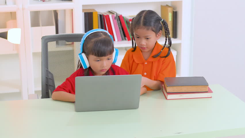 Two young asian girls collaborating on a laptop for online education, wearing headphones and glasses, with books stacked nearby, embodying remote learning and teamwork - Powered by Shutterstock - Get 15% off with code: PIKWIZARD15