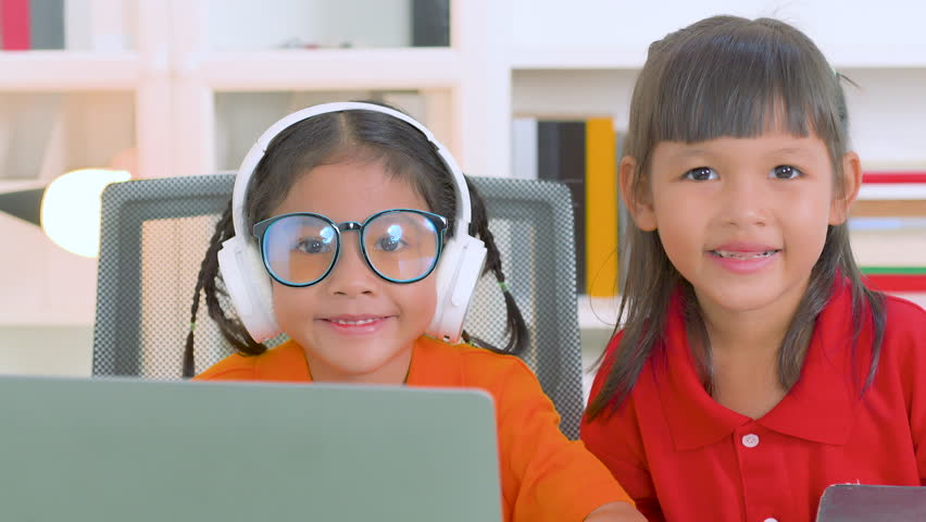 Two young asian girls collaborating on a laptop for online education, wearing headphones and glasses, with books stacked nearby, embodying remote learning and teamwork - Powered by Shutterstock - Get 15% off with code: PIKWIZARD15