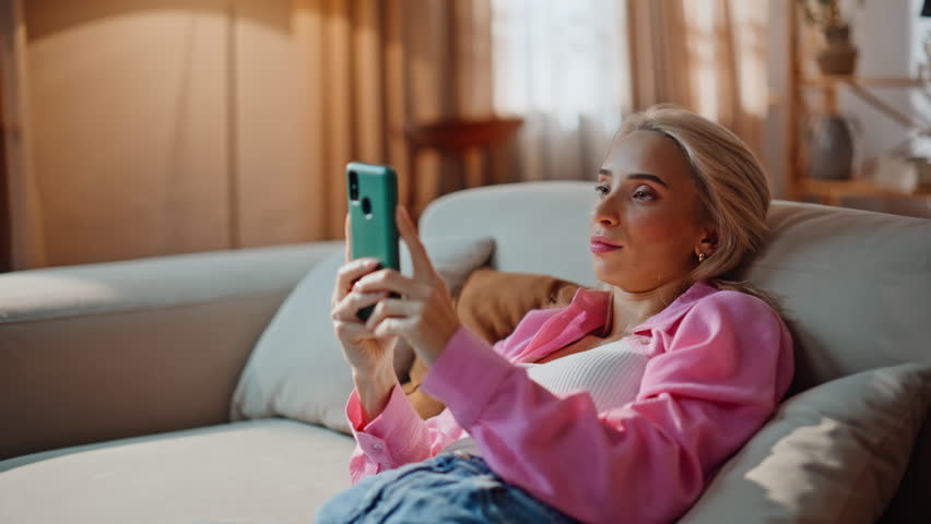 Relaxed blonde staring mobile phone lying at home sofa closeup. Chilling woman shopping remotely by smartphone at domestic weekend alone. Attractive girl thinking on online purchase looking cellphone