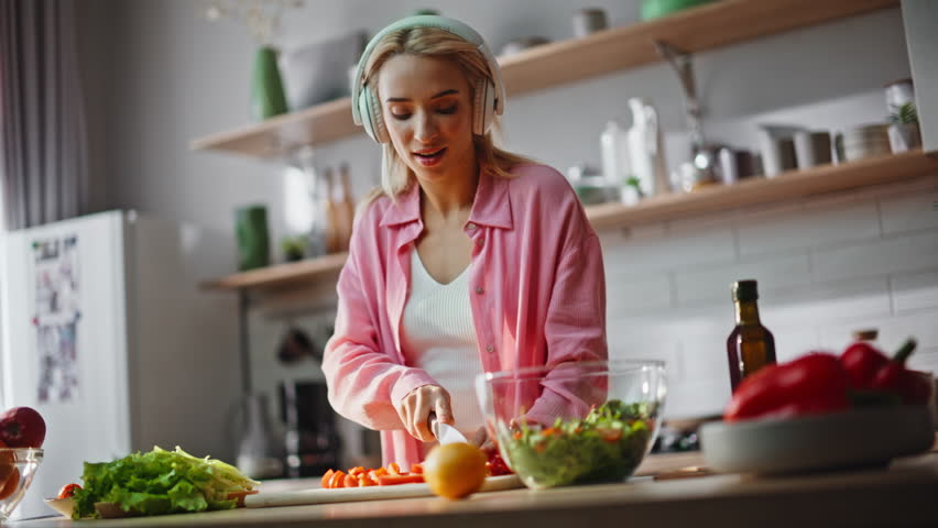 Girl preparing food in headphones singing along favorite song at cozy kitchen. Carefree joyful woman dancing listening music in headset cooking salad closeup. Smiling blonde chopping vegetables alone 