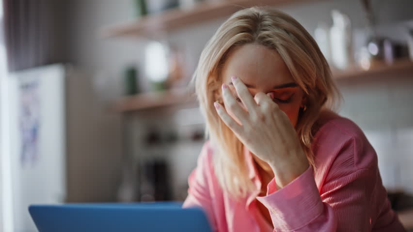 Exhausted blonde rubbing neck experiencing fatigue at kitchen portrait. Frustrated businesswoman working laptop closeup. Overworked woman tired from deadline sitting at light apartment interior alone 