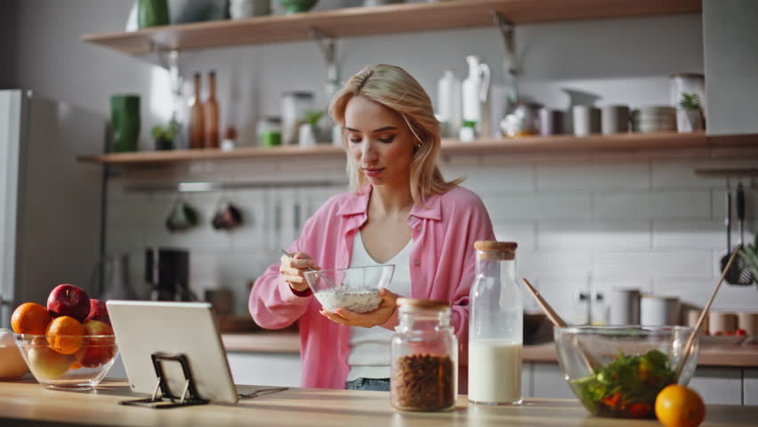 Online woman having breakfast looking tablet in apartment kitchen closeup. Attractive lady in pink shirt eating granola watching movie on pad computer. Relaxed girl enjoying meal in light cuisine 