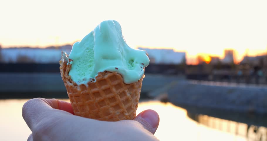 Delicate hand holding melting pistachio ice cream in waffle cone, highlighting sunset river scenery with golden evening light and soft color reflections