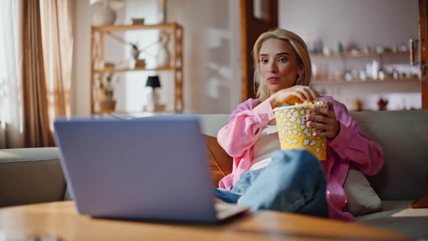 Emotional woman watching interesting film eating popcorn at sunny apartment zoom on. Focused girl engaged in intense moment movie at home couch. Laughing lady enjoy funny tv show having fun on sofa