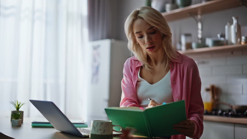 Smart girl freelancer making notes in notepad looking laptop screen at kitchen closeup. Thoughtful woman working computer sitting cozy modern kitchen. Focused businesswoman watching online webinar
