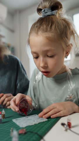 Girl pours red UV resin into silicone mold, making small figures under adult supervision. Safe polymer experiments for kids at home, bonding time with family, developing manual skills.
