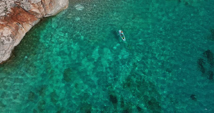 Aerial parallax of person on SUP board in turquoise water by cliff