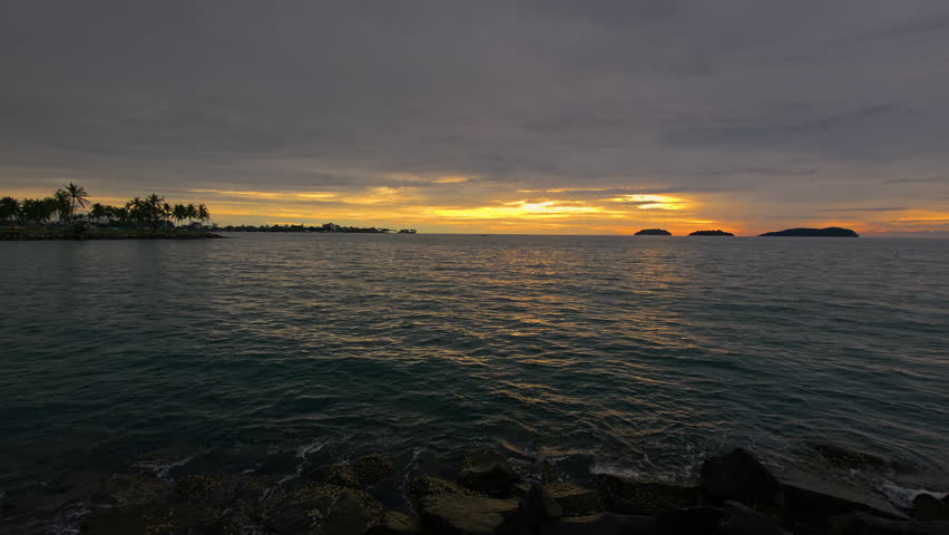 Kota Kinabalu, Sabah, Malaysia -
Golden Hues Across Cloudy Sunset Sky Over The Sea, Wave Crashing Against Rocky Beach