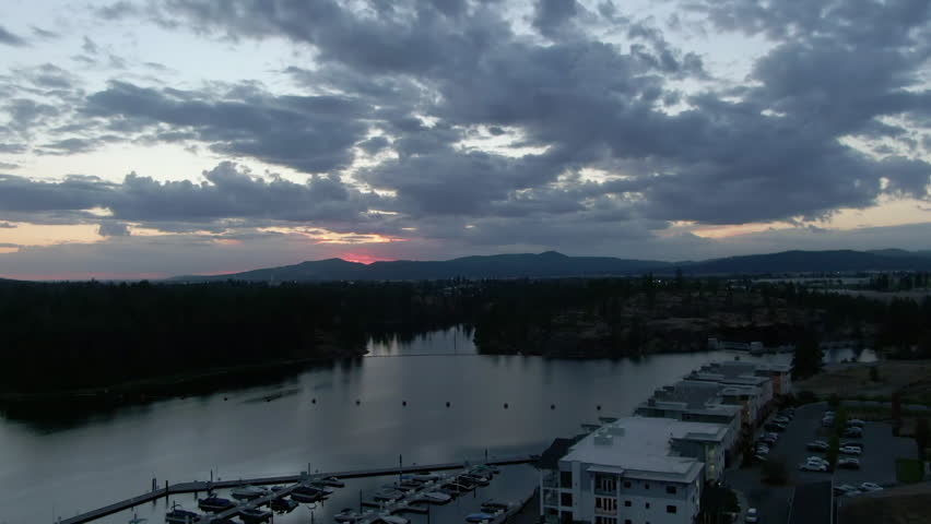 Aerial Shot Of Nautical Vessels Moored At Harbor In Lake By Mountains Under Cloudy Sky, Drone Flying Backwards During Sunset - Coeur d`Alene, Idaho