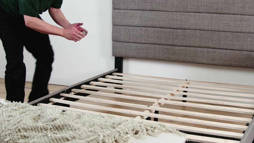 Man documents bed slat condition from several angles in wood-floored bedroom