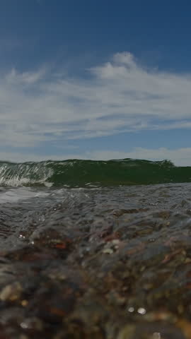 Vertical video, Close-up from the lower level, Wave after wave runs onto the pebble shore covering the camera, underwater forming air bubbles, Underwater view, Slow motion