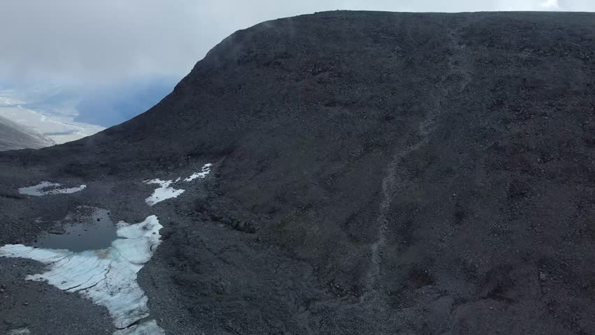 Aerial drone footage view of Large black colored Kebnekaise Volcanic terrain mountain during a cold chilly day with white snow patches on the mountain. Hiking trail leading up the mountain, cloudy day