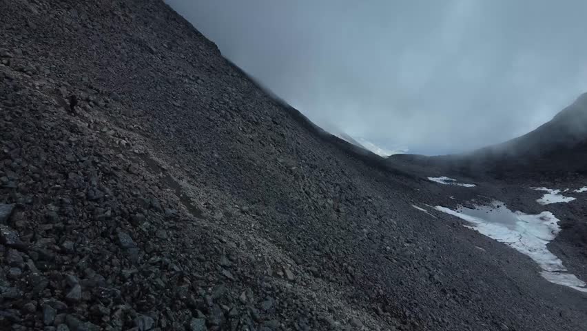 Aerial drone footage following a person hiking up a small trail in dark colored volcanic terrain Kebnekaise mountain in Sweden whil white and blue clouds are low to the ground visible in the back.
