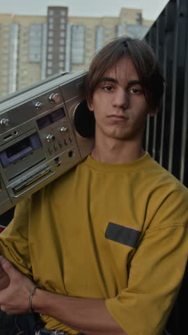 Vertical medium portrait of Caucasian teenage boy in streetwear looking at camera standing outdoors and holding silver old school cassette boombox on shoulders