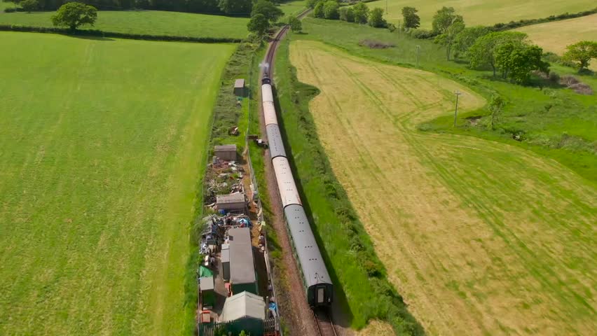 Slow motion aerial of a steam train gliding through open farmland near Swanage, Dorset, with green pastures, sheds, and scenic rural landscape