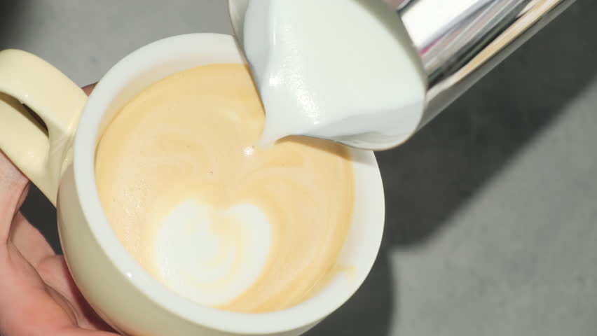 Woman carefully adds milk to coffee forming design on surface. Barista demonstrates final step crafting latte showcasing working process