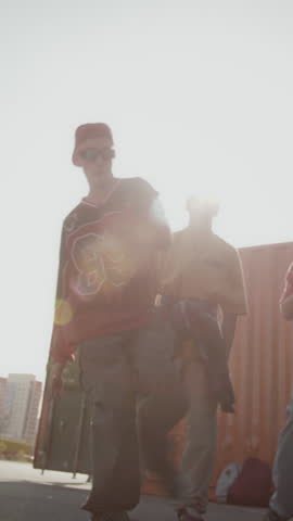 Vertical handheld shot of talented Caucasian teenager performing breakdance in front of peers supporting him on bright summer day outdoors