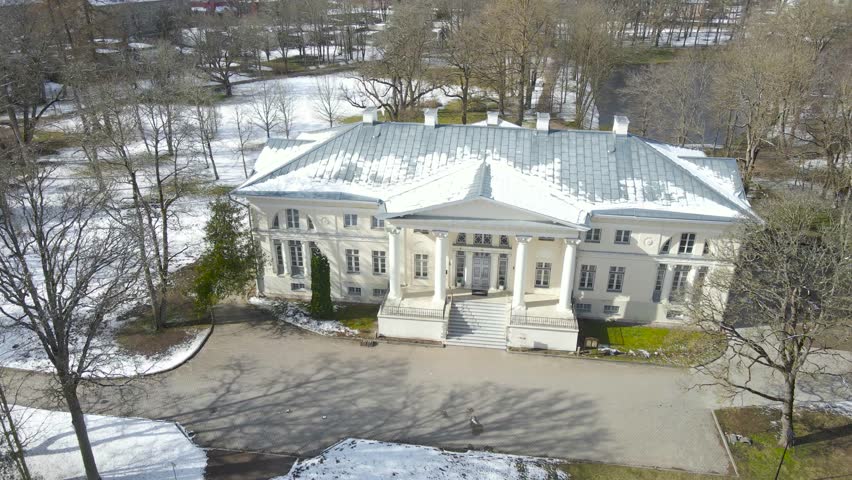 Aerial drone footage orbiting and spinning around a gorgeous large white mansion or manor called Saku mõis during winter sunny day while green mowed lawn grass visible in the garden and leafless trees