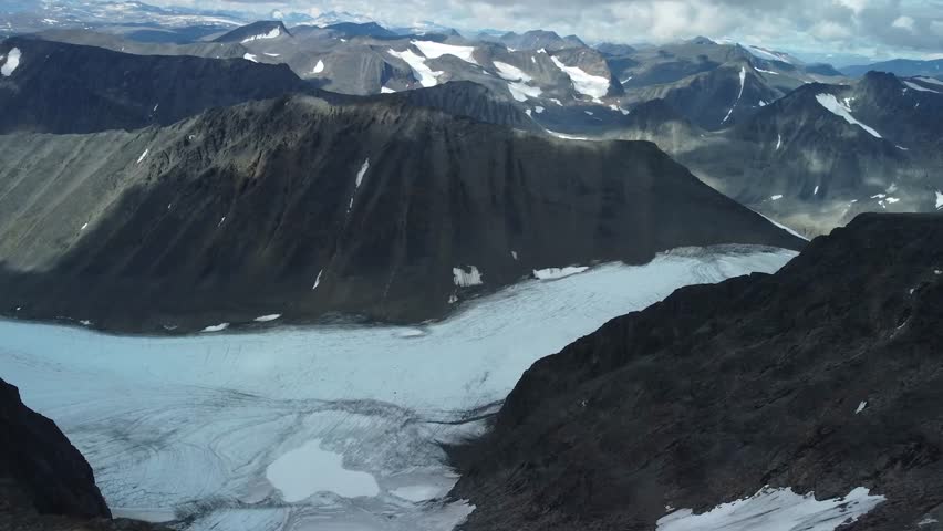 Aerial drone footage slowly panning down and revealing a white snow covered snowy landscape between tall and steep black colored Kebnekaise mountains during a sunny and cloudy day in Sweden noric land