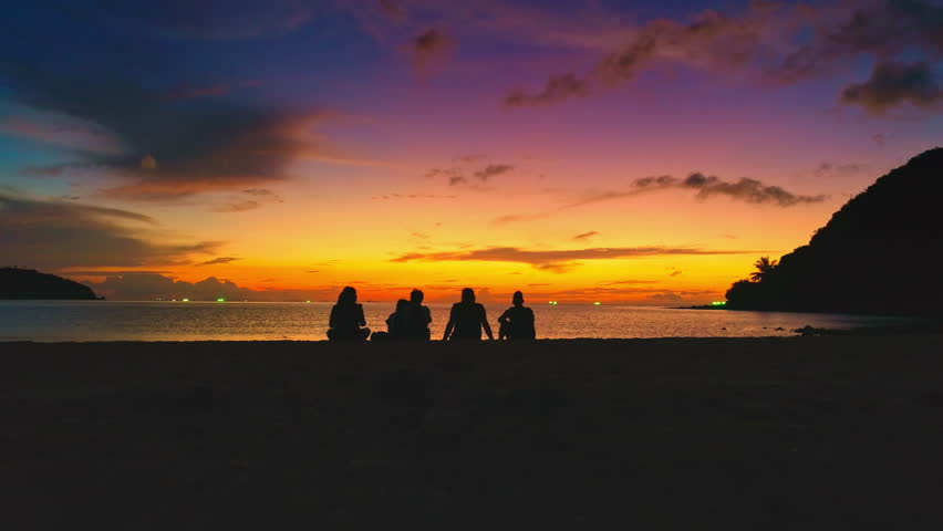 Five tourists sitting on the sandy shores of Koh Ma Beach, Koh Phangan Island, Thailand, are admiring a stunningly colorful sunset painting the sky over the tranquil sea