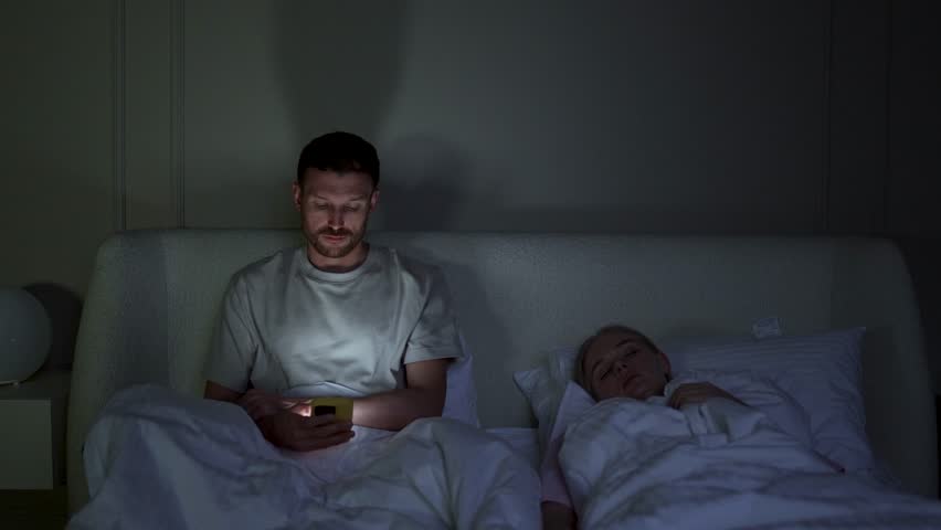 Stressed man engrossed in smartphone at night, disturbing wife sleep in bed. Male struggle with insomnia, phone digital addiction, endless scrolling through social media, sleep disruption, nomophobia