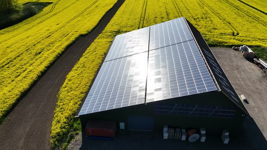 Aerial footage of large solar panel system on the roof of a modern agricultural barn, surrounded by yellow rapeseed fields in spring. Farm equipment storage with large solar photovoltaic  array. 