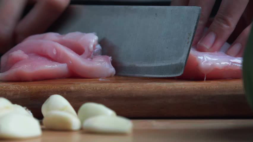 A woman is cutting chicken breast fillet into strips on a wooden cutting board