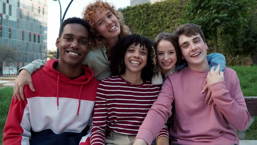 Happy multi-ethnic friends smiling in slow motion. Four cheerful multi-ethnic young students smiling and hugging, sitting on bench in university campus