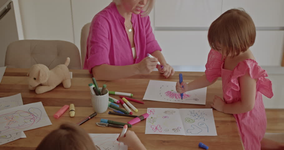 A mother and her two daughters enjoy quality time together, drawing and coloring at the kitchen table.