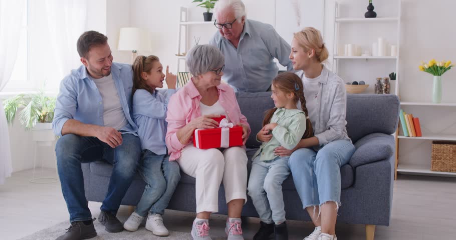 Happy cheerful grandchildren with parents giving present gift boxes to their senior elderly grandmother sitting on sofa at home congratulating her on holiday. Love and family concept. 4k video.