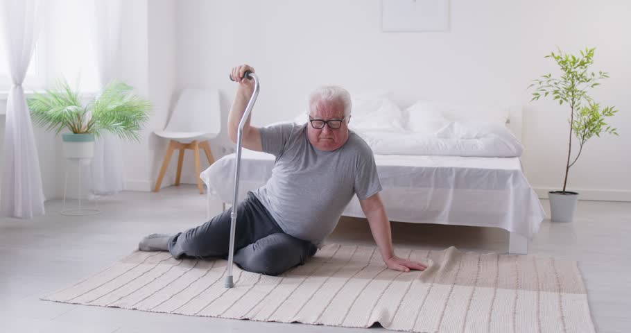 Unhealthy senior man stands up from the floor after a fall caused by an accident or health problem at home. Man struggles with injury or disease, highlighting the dangers of trauma for elderly people.
