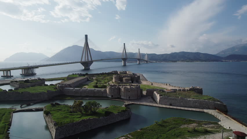 Stunning drone footage of the famous Rio-Antirrio Bridge spanning the Gulf of Corinth, with historical ruins in the foreground.