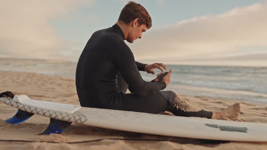 A surfer in a wetsuit sits on his surfboard on a sandy beach and checks his social media between sets.