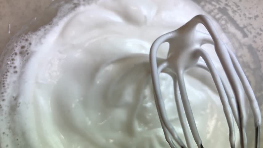 A mixer whips sugar and eggs in a glass bowl in the kitchen. Beat the meringue from egg whites. Top view. 