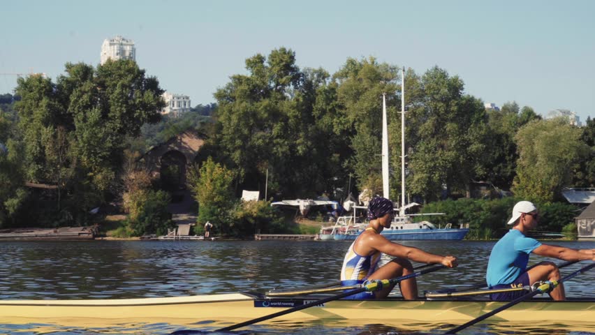 Rowing team training. Side view of 2 young caucasian male rowers, during a rowing practice, athlete sitting in a boat in the river Dnipro, rows through a calm water sunny day, autumn. 4k footage