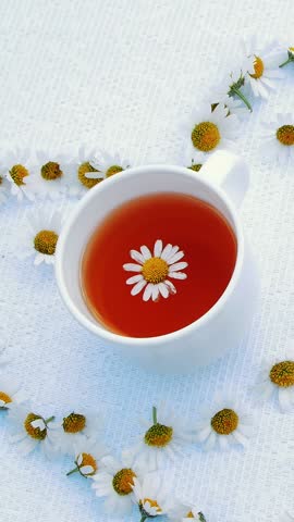 A white plate with pastries, biscuits and cookies, decorated with white daisies. On white saucer there is one white cup with chamomile tea. Around It, there is a heart of chamomiles