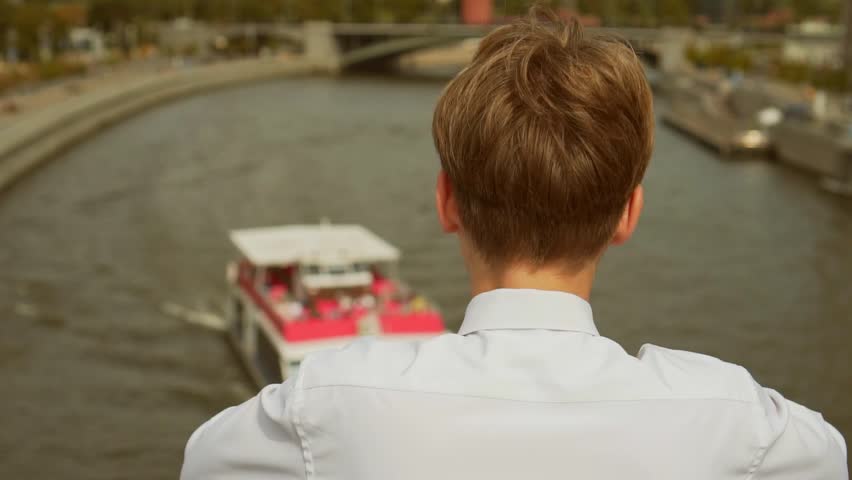 A young man stands on a bridge and looks at a boat passing on the river